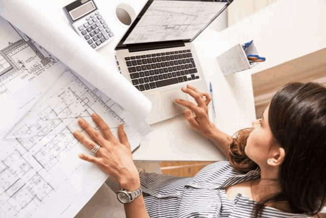 a woman sits at a desk looking at a laptop and a calculator