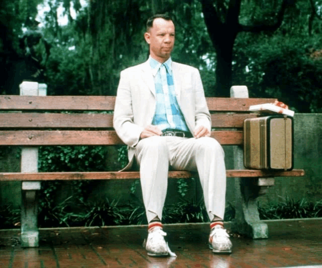 a man in a suit sits on a park bench with a suitcase on his lap