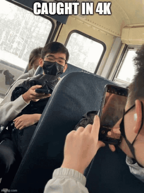 a man wearing a mask takes a picture of another man on a bus with the caption " caught in 4k "