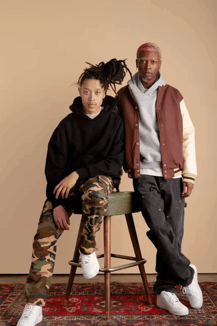 a man in a varsity jacket sits next to another man in camo pants