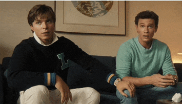 two men are sitting on a couch with one wearing a sweater with the letter i on it