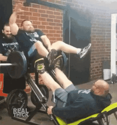 a man is laying on a bench while another man lifts a barbell over his head in a gym .