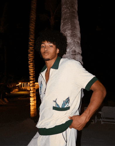 a man wearing a white shirt with a green collar is standing next to a palm tree