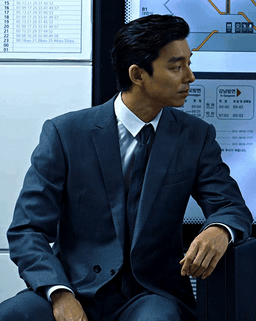 a man in a suit and tie is sitting in front of a sign that says ' b1 ' on it