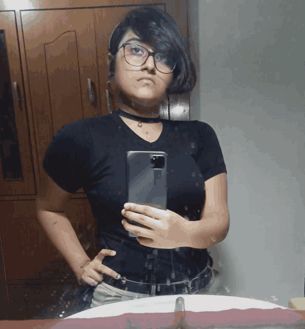 a girl wearing glasses and a choker takes a selfie in front of a mirror