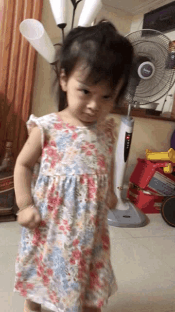 a little girl in a floral dress stands in front of a fan that says ' panasonic ' on it