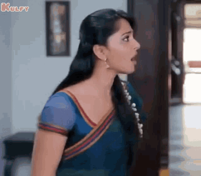 a woman in a blue saree is standing in a room and making a funny face .