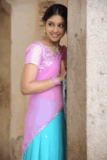 a woman in a pink and blue saree is standing next to a stone wall .