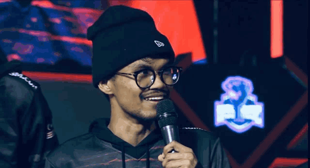 a man wearing glasses and a new era hat holds a microphone