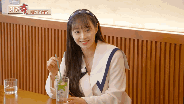 a woman sits at a table with a glass of water and a straw in front of a sign that says " 지켜 "