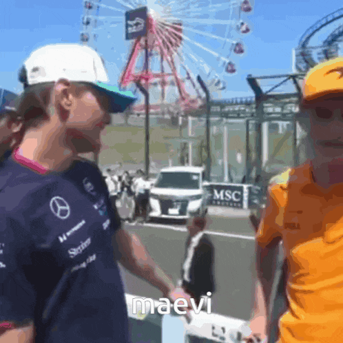 a man in a mercedes shirt shakes hands with another man in an orange shirt in front of a ferris wheel