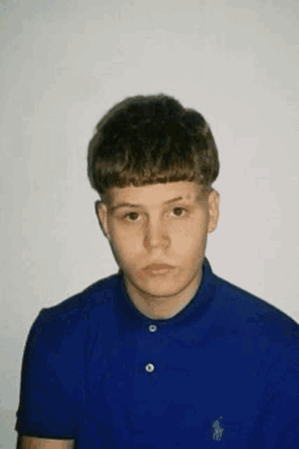 a young man wearing a blue polo shirt is standing in front of a white wall and looking at the camera .