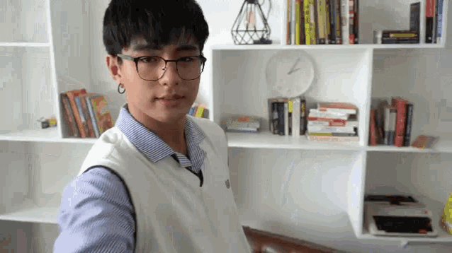 a young man wearing glasses and a vest is standing in front of a bookshelf with books on it .