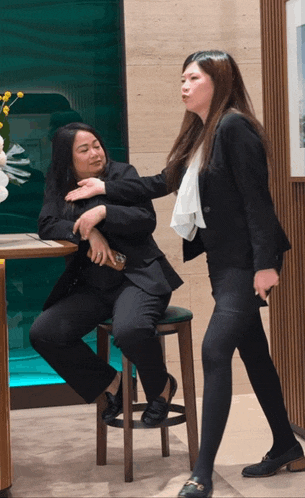 a woman in a suit sits on a stool next to another woman in a suit