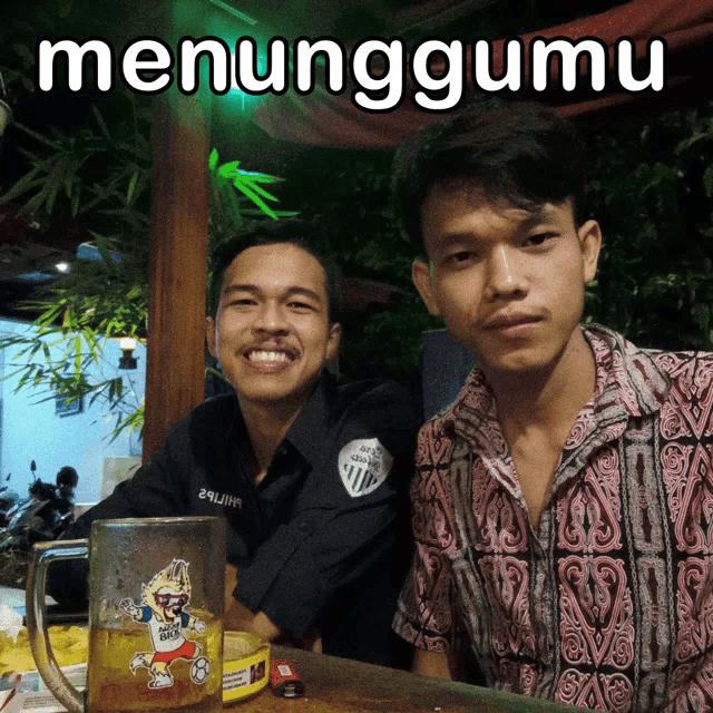 two men are sitting at a table with a mug of beer and the word menunggumu on the bottom