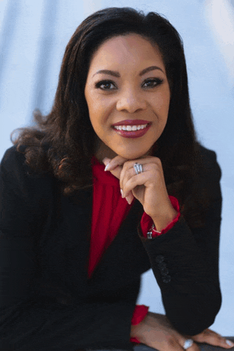 a woman wearing a black jacket and a red shirt smiles with her hand resting on her chin