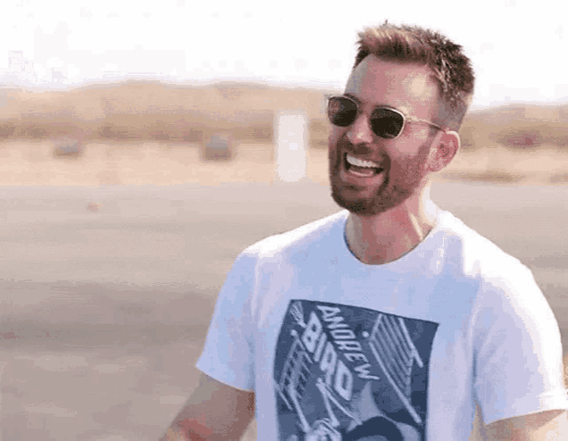 a man with a beard wearing sunglasses and a white t-shirt is smiling .