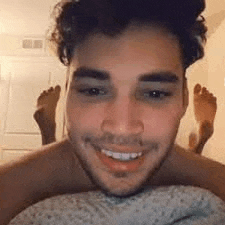 a man is smiling while laying in bed with his feet up .