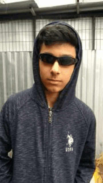 a young man wearing sunglasses and a hoodie is standing in front of a metal fence .