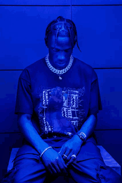 a man wearing a t-shirt that says ' travis scott ' on it is sitting in front of a blue wall