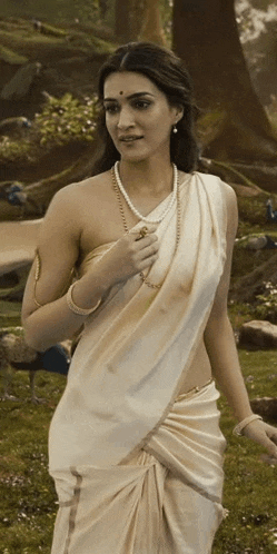 a woman wearing a white saree and pearls is standing in a field .