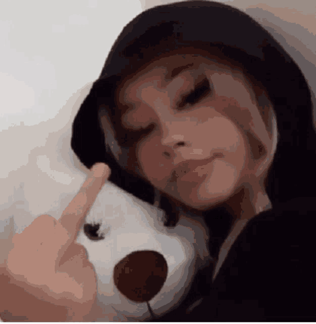 a girl in a hoodie is holding a teddy bear and giving the middle finger .