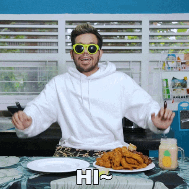 a man wearing sunglasses and a white hoodie is sitting at a table with a plate of food and a sign that says hi