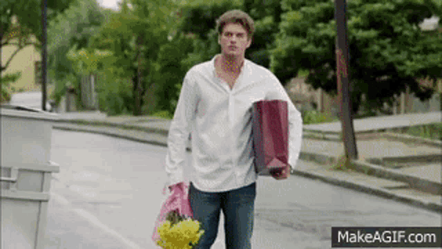 a man in a white shirt is walking down the street holding a bag of flowers and a gift bag .