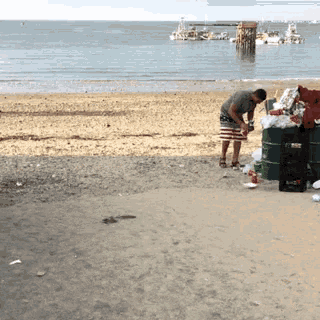 a man is standing on a beach next to a green barrel that says ' coca cola ' on it