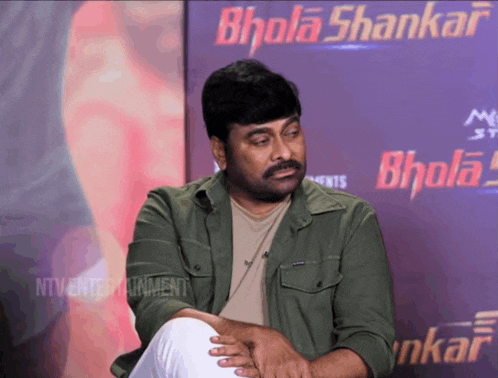 a man sits in front of a bhola shankar poster