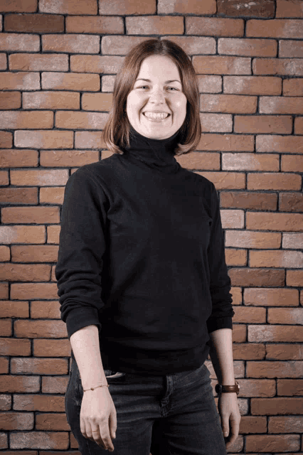 a woman wearing a black turtleneck sweater smiles in front of a brick wall