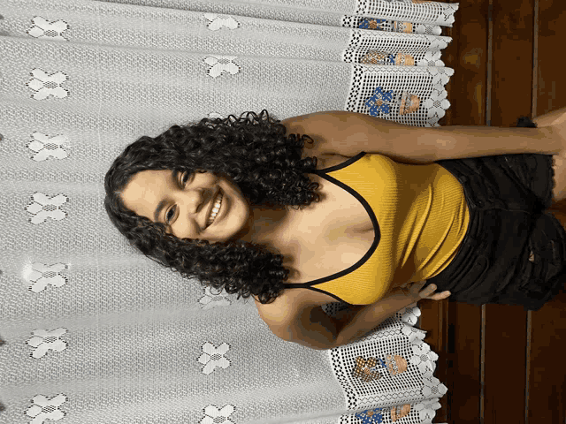 a woman in a yellow tank top is smiling in front of a white curtain with flowers on it