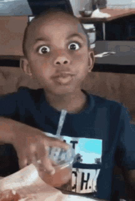 a young boy is sitting at a table drinking from a plastic cup with a straw and making a funny face .