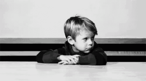 a black and white photo of a young boy sitting at a table with his hands on his face .