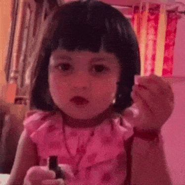 a little girl in a pink dress is holding a bottle of nail polish and making a funny face .