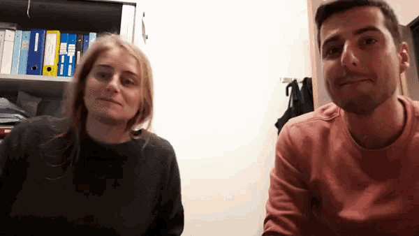 a man and a woman are sitting next to each other in front of a shelf with binders on it