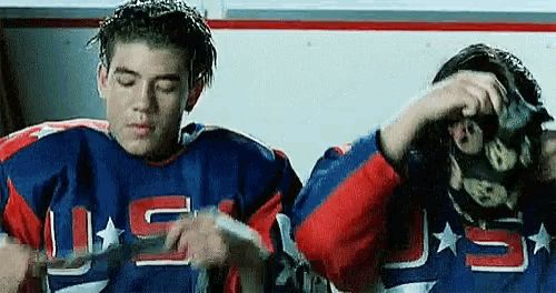 two hockey players are sitting next to each other in a locker room . one of the players is wearing a mask .