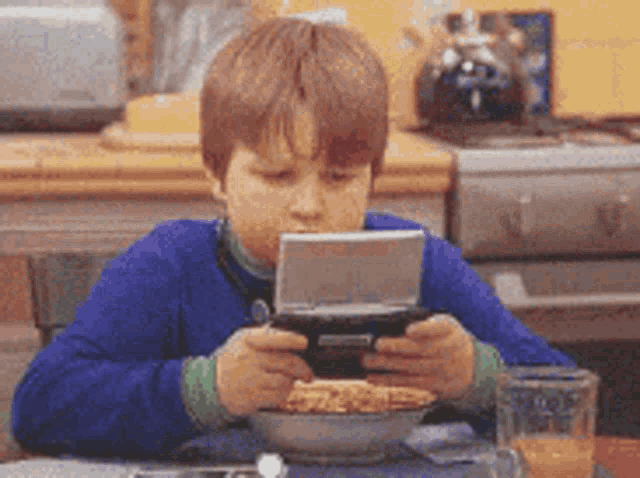 a young boy sitting at a table with a bowl of cereal and a glass of orange juice playing a video game