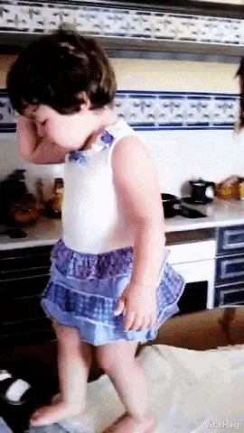 a little girl in a blue and white dress is standing in a kitchen and covering her face with her hands