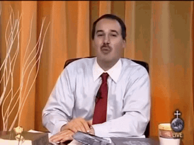 a man in a white shirt and red tie is sitting at a desk in front of a curtain .