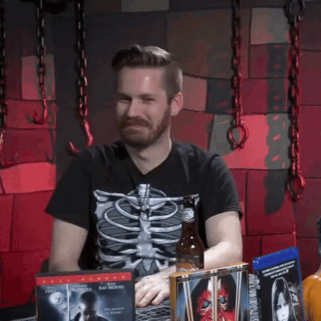 a man wearing a skeleton shirt is sitting at a table with a bottle of beer and some dvds .