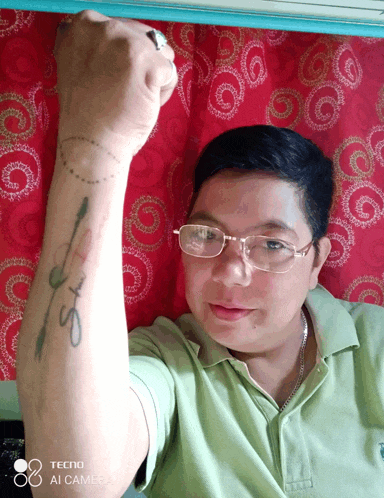 a man wearing glasses and a green shirt has a tattoo on his arm that says ' s ' on it