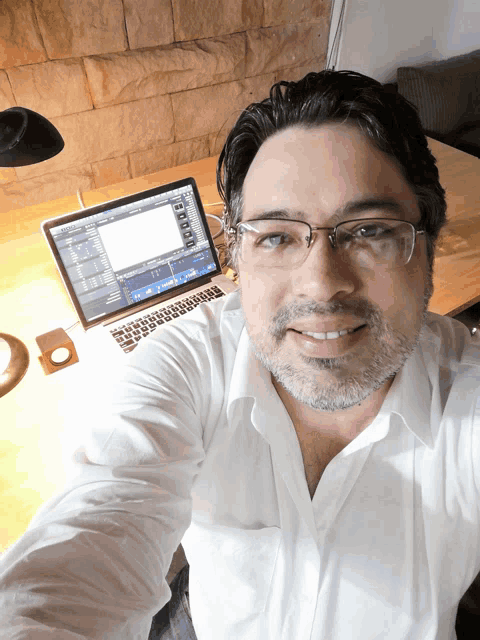 a man wearing glasses takes a selfie in front of a macbook