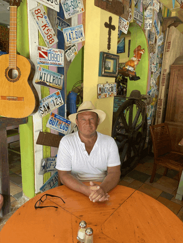 a man wearing a white shirt and a hat sits at a table in front of a wall of license plates