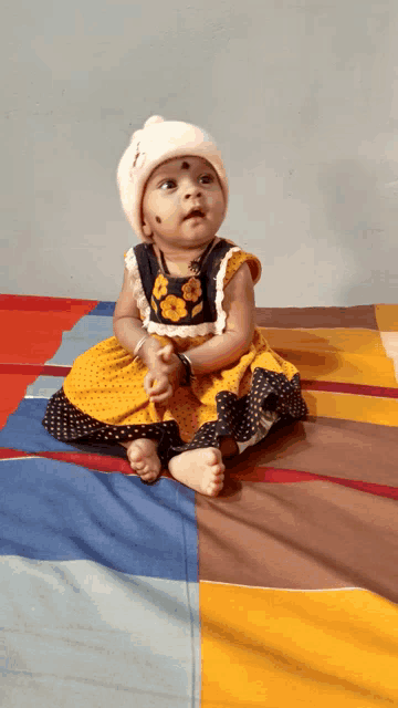 a baby wearing a yellow and black dress and a white hat sits on a bed