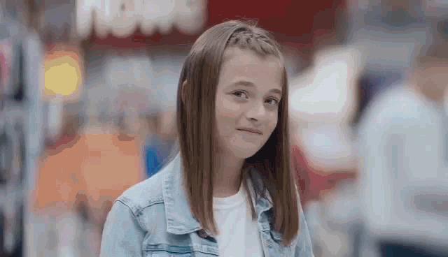 a young girl in a denim jacket and white shirt is standing in a store and looking at the camera .