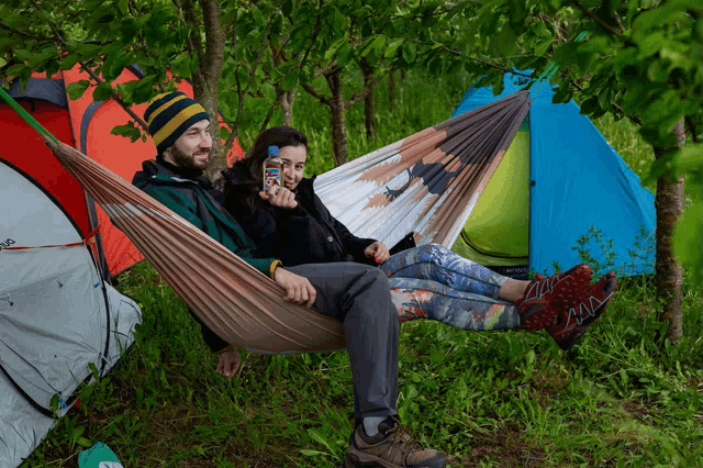 a man and a woman are sitting in a hammock with tents in the background and the man is holding a bottle of water