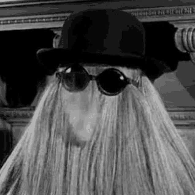 a black and white photo of a person wearing a hat , sunglasses and a wig .