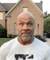 a bald man with a beard is wearing a white shirt in front of a brick house .