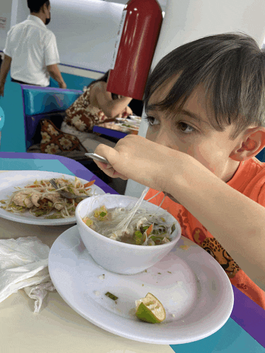 a young boy in an orange shirt is eating a bowl of soup with a spoon